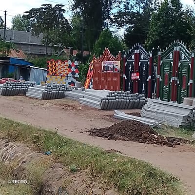 This image depicts an outdoor commercial display of construction materials and gates located in Mumbuni North ward, Kenya. The foreground consists of a dirt path, bordered on the left by a narrow grassy strip and a shallow ditch. A pile of dark brown soil is situated on the path in the mid-right foreground.
Along the midground, numerous stacks of grey concrete fence posts or pillars are arranged horizontally. To their left, a blue corrugated metal structure is partially visible, with an individual partially obscured, appearing seated nearby. Adjacent to the concrete posts, stacks of colorful interlocking paving blocks displaying patterns in yellow, grey, black, orange, and red are present. Further right, a series of ornate metal gates are displayed upright. The prominent central gate is reddish-brown, featuring an elaborate top and a sign that reads "BRIGHTON CAIRO WORKS" along with partial contact numbers. To its right, two additional ornate metal gates are exhibited: one predominantly black and red, and the other green and red, both equipped with decorative spikes or finials. More stacks of grey concrete elements, resembling curbing or additional posts, are interspersed among the gate displays.
In the background, various buildings with corrugated metal roofs and grey block walls are visible, consistent with a residential or commercial area. These structures are partially obscured by dense green foliage from numerous trees, visible under a partly cloudy sky.