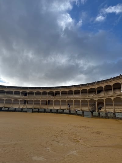 The image displays an interior view of the Plaza de Toros, a bullring located in Ronda, Spain. The scene is dominated by a circular, sandy arena floor, which appears largely empty, with visible darker, possibly damp, patches. Encircling the arena is a low, decorative barrier constructed of grey and white tiles. Beyond this barrier, the bullring's structure rises with multiple tiers of arcaded seating, built from light-colored stone or concrete. The structure features a lower seating area, topped by two distinct levels, each comprising a series of uniform arches supported by columns. The uppermost edge of the bullring is adorned with decorative architectural details.

On the right side of the image, a grey metal gate is visible within the barrier, providing access to the seating areas or internal corridors. A few individuals are present on the lower seating tier: one person stands near the barrier, seen from the back, dressed in dark clothing, while two other figures are partially visible, seated or standing further back in the seating area. The sky above is largely overcast with extensive dark grey clouds, though patches of clear blue sky are visible in the upper right portion of the frame.