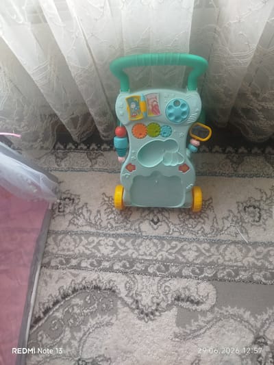 The image displays a mint green baby walker with various colorful interactive toys, including gears, buttons, and a small mirror. The walker is positioned on a patterned carpet, which appears to be in a living room. Behind the walker, sheer white curtains are visible, suggesting a window. The lighting is soft, indicating it might be daytime. The bottom left corner of the image shows "REDMI Note 13" and the bottom right corner displays "29.06.2026 12:57," indicating the device and date the photograph was taken. No people are present in the image. The image is not taken in Tokmok City, Kyrgyzstan.