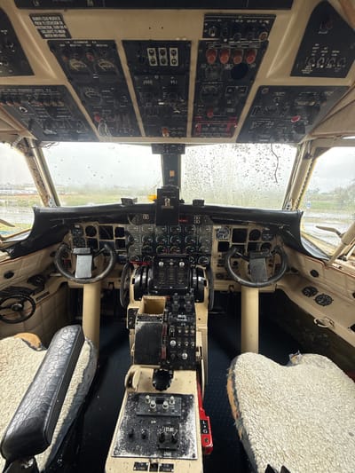 This is a photograph of the interior of an aircraft cockpit. The cockpit features a complex array of instruments, gauges, and control panels. Two pilot seats covered in a shaggy, light-colored material are visible. Through the front windshield, it is raining. The control panels display labels such as "LH ALTERNATOR," "RH MECH FAIL," "DE-ICE SEQUENCE LIGHTS RH," and "DME." The text "ZS-LYL" is also visible. There is no specific information available to pinpoint the location in Kraaifontein, South Africa. However, it can be implied to be at an aircraft museum or a similar facility.