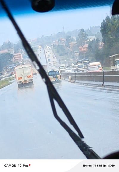 The image depicts a rainy daytime scene on a multi-lane highway in Muguga ward, Kenya, viewed from the interior of a vehicle through its windshield. A dark, segmented windshield wiper blade occupies the lower central and right portions of the frame, angled upwards to the left. The road surface is uniformly wet and reflective, with visible water spray originating from moving vehicles. Traffic on the highway is moderate to heavy, comprising large white commercial box trucks, smaller white vans or pickup trucks, and various passenger vehicles, moving in both directions. A concrete median barrier separates the highway lanes. Positioned on the right side of this median barrier, adjacent to the closest lane, are at least two tan-colored statues resembling crouching lions. The background features a hilly, urbanized landscape with numerous multi-story residential buildings, predominantly in earth tones (orange, brown, beige), interspersed with dense green vegetation and trees, all under an overcast sky. A tall, slender structure, possibly a communication tower, is visible on the distant horizon. No individual persons are distinctly visible outside of vehicles.