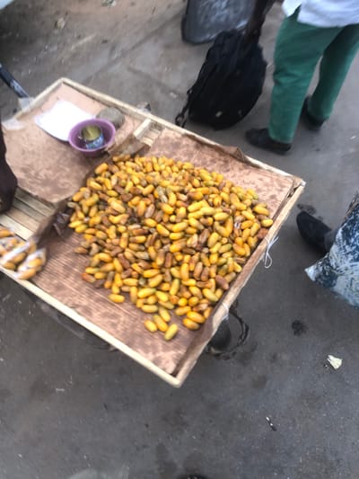 The scene depicts a street-level vendor's display in Tukur Tukur, Nigeria. A rectangular wooden frame, approximately 1 meter in width, serves as a display counter, covered with a brown, possibly cardboard, liner. Piled centrally on this surface is a large quantity of ripe, yellow to yellowish-brown, oval-shaped fruits, resembling dates. To the immediate left of the main fruit pile, a smaller quantity of the same fruit is visible, contained within clear plastic packaging. On the upper-left section of the display, a small purple plastic bowl holds a metallic can or cup, with a piece of white paper situated nearby. The ground is dark grey, appearing to be asphalt or concrete, with scattered dirt and small debris. Three individuals are partially visible: a person's left foot and lower leg in a dark sandal are present on the left side of the frame; another person, wearing green trousers and dark closed-toe shoes, stands near the right side of the display, with a black backpack resting on the ground behind their legs; and the lower legs and feet of a third person, dressed in a patterned garment and dark closed-toe shoes, are visible on the bottom right.