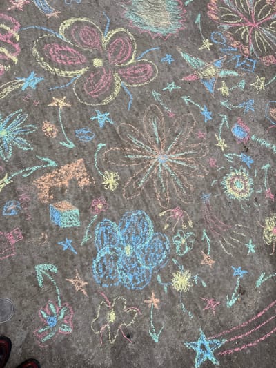 The scene depicts a dark grey, textured pavement surface covered extensively with numerous colorful chalk drawings. The drawings are primarily in pastel shades of pink, yellow, light blue, teal, and orange. Dominant motifs include various styles of multi-petaled flowers, five-pointed stars, some with trailing lines, arrows, and abstract squiggles. Specific identifiable shapes include a light blue cube and an orange house-like structure. The drawings are densely distributed across the entire visible area, with some overlapping. In the bottom left corner, the tips of two feet in dark footwear are partially visible, one with a grey sole and the other with a dark red/brown upper. The location is Saint-Mandé, France.
