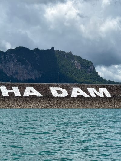 This media file shows a vertical view of a dam. The dam wall is constructed from dark-colored rocks and features large, white lettering that reads "HAD YAI DAM". To the left of the dam, a lush green mountain rises, partially obscured by a cloudy sky. The water on the right side of the dam is a deep blue-green, with ripples indicating a gentle surface movement.

Several people are visible on the dam, scattered along its length, suggesting it is open to visitors. Some appear to be standing, looking out at the water or the landscape. A few small poles, possibly lights or flagpoles, are also present on the dam.

The weather appears overcast, with a mix of dark and lighter clouds in the sky. The overall scene suggests a daytime setting, possibly late afternoon given the diffused lighting. The location is identified as Ban Chiao Lan, Thailand, which is known for its picturesque dam and surrounding natural beauty. The image evokes a sense of natural grandeur and human engineering, with the dam acting as a boundary between the rugged mountains and the expanse of water.
