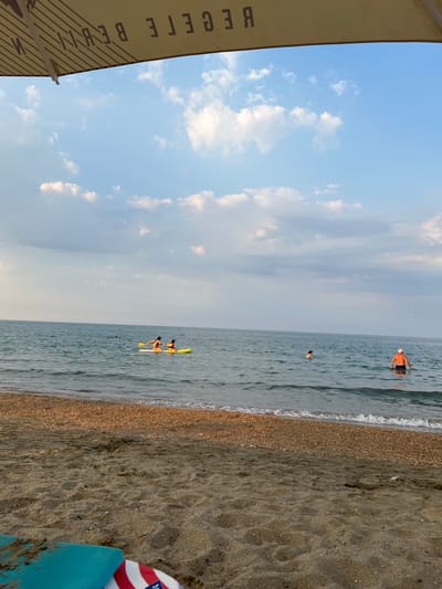 The image depicts a beach scene with a focus on the ocean and shoreline. Several people are visible in the water. In the mid-distance, two individuals are on a yellow paddleboard, with one person holding a paddle. Further out, a person in an orange top is wading in the water, and another person is also in the water in the distance. The sea is calm, with gentle waves lapping the shore. The beach itself is composed of sand and small pebbles. The sky is partly cloudy with a pale blue hue, suggesting it is either early morning or late afternoon. A portion of a canvas umbrella with diagonal lines and some text is visible in the lower left corner of the frame, partially obscuring the sky. The text visible on the umbrella appears to be "BEGETE BEY." The overall mood is relaxed and leisurely, indicative of a day at the beach. The location is Constanța, Romania.