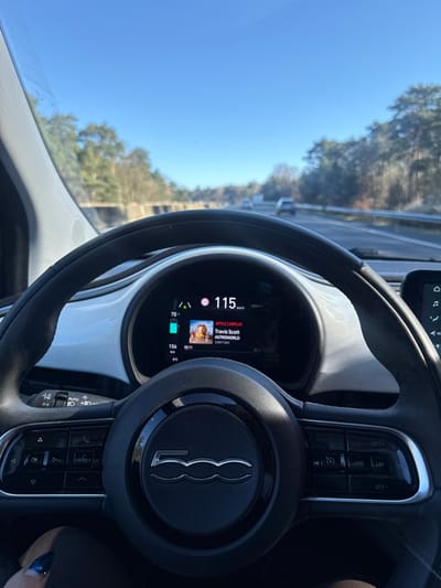 The image displays the interior of a Fiat 500 vehicle from the driver's perspective, traveling on a multi-lane highway near Thiers-sur-Thève, France, under a clear blue sky. The black, leather-wrapped steering wheel, featuring a chrome "500" emblem at its center, dominates the lower half of the frame. A hand with dark nail polish is visible to the lower left of the steering wheel. The digital instrument cluster behind the steering wheel shows the vehicle's speed at 115 km/h, with a detected speed limit of 70 km/h displayed. The battery charge is at approximately 70%, indicating a range of 154 km. Apple CarPlay is active, displaying the album "ASTROWORLD" by Travis Scott and the song "Can't Say." The external temperature is 8°C. Through the windshield, the highway extends into the distance with several vehicles ahead in receding lanes. The road is bordered by a concrete barrier to the left and a metal guardrail to the right, beyond which dense tree lines are visible. The scene is illuminated by bright sunlight.