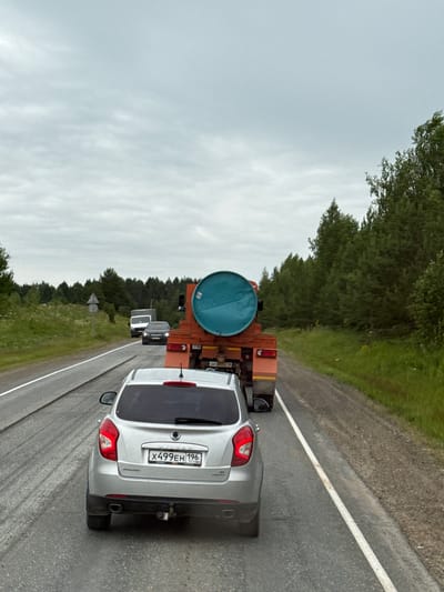 This is a photograph taken from a vehicle looking ahead on a rural road. The sky is overcast. In the foreground, a silver SUV is stopped on the right side of the road. The SUV has a license plate that reads "X499EH 196". Behind the SUV, an orange truck with a large blue barrel strapped to its flatbed is visible. The truck is also stopped on the road. The road is paved and has a white line marking the lane. On the left side of the road, there is a grassy embankment with trees in the distance. On the right side, there is a gravel shoulder and more trees. The overall scene suggests a traffic delay. The location is specified as Гремяча, Russia.