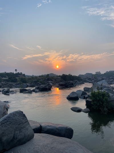 This image depicts a wide view of a rocky river landscape in Hale Bandi Harlapura, India, under the conditions of either sunrise or sunset. The sky, occupying the upper portion of the frame, transitions from a pale blue at the zenith to an orange-yellow hue near the horizon, with scattered wispy clouds visible. A distinct, bright orange-yellow sun disc is positioned centrally above the horizon, casting its light across the scene.
A river flows horizontally through the midground, its surface largely calm with visible reflections of the sun's direct light as an elongated orange streak, and the general sky colors. Numerous dark grey to black boulders of varying sizes are present within the river and along its banks. In the foreground, several large, dark boulders are visible, partially submerged on the bottom left and bottom center. The riverbanks on both sides are characterized by a dense scattering of these dark, rounded rocks. Sparse green vegetation, primarily bushes and small trees, grows among the rocks along the banks. A tall, slender, single-trunked tree, resembling a palm, is silhouetted against the sky on the left bank. In the distant background, low-lying, rocky landforms with minimal vegetation are visible. No individuals, animals, or human activities are observed within the frame.