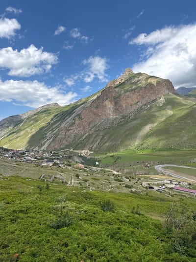 A panoramic, eye-level view captures the picturesque mountain village of El'tiubiu, Russia, nestled at the foot of a colossal, verdant mountain under a bright blue sky dotted with fluffy white clouds. The foreground is dominated by a lush expanse of green, low-lying vegetation, creating a soft, textured carpet.

The middle ground reveals the village, a collection of buildings with diverse roof colors, including red, white, and gray, spread across a gentle slope. Some houses feature traditional architectural elements, while others appear more modern. A winding road, visible in the mid-right, curves through the landscape, with several vehicles parked along its side near a building with a red roof. A small river snakes its way through the valley, adding a touch of serenity to the scene. The terrain is varied, with patches of grass, scattered rocks, and evidence of human activity such as pathways and stone ruins.

The majestic mountain takes center stage in the background, its steep slopes covered in vibrant green grass, interspersed with exposed rocky faces displaying shades of brown and tan. The sunlight casts dramatic shadows across the mountain, highlighting its rugged contours and creating a striking play of light and shade. The peak of the mountain is sharp and prominent, reaching towards the expansive sky. The overall impression is one of natural grandeur and tranquil rural life.