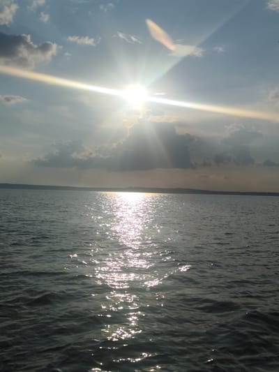 The image depicts a vast body of water, likely a lake or sea, under a cloudy sky illuminated by the sun. Sunbeams pierce through the clouds, creating bright streaks of light that reflect off the rippling surface of the water, forming a shimmering path. The horizon line is barely visible, suggesting a wide expanse of water. The water itself appears dark and slightly choppy. The sky is a mix of blue and grey clouds, with the sun positioned high in the frame. No people or man-made structures are visible in the image. The location of Галево, Russia is not discernible from the visual information provided in the image.