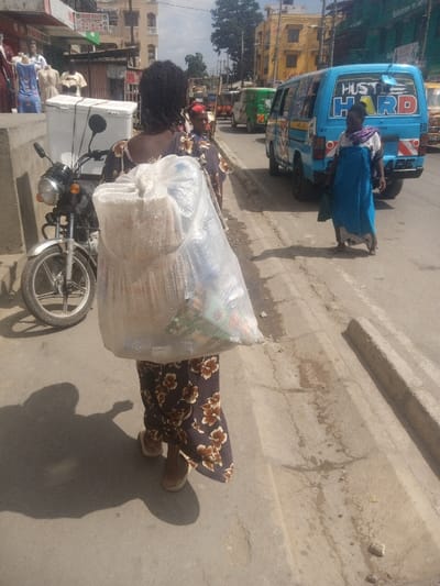 A woman carries a large plastic bag filled with goods while walking along a street. The bag appears heavy and bulky. A parked motorcycle with a white box attached is visible on the left. Another woman in a blue cloth is walking away from the camera. A blue van with "HUSTLE HARD" written on its back is also visible. Other vehicles are parked on the street. The street is concrete and shows signs of wear and tear.