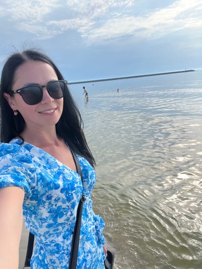 A woman with dark hair, wearing sunglasses and a blue floral dress, takes a selfie at the water's edge in Baltiysk, Russia. She stands on a sandy or rocky shore, with calm water stretching out before her. In the distance, a long stone breakwater extends into the sea, with a lighthouse at its end. Several people are visible in the water, wading or swimming. The sky is partly cloudy, with a bright sun reflecting off the water's surface. The overall scene suggests a pleasant day at the beach.