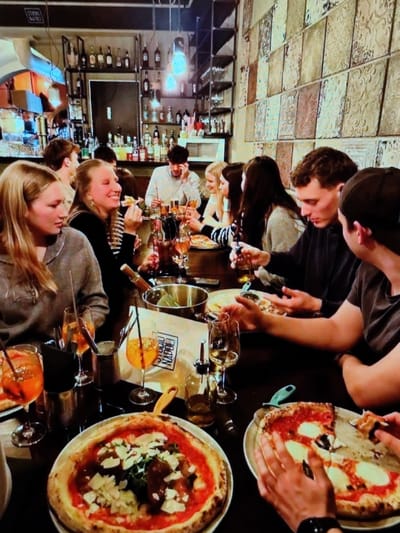 The image depicts an interior scene at "Studio Napoli," a restaurant located at Place du Centenaire 17, 1420 Braine-l'Alleud, Belgium. Approximately ten young adults are seated around a dark wooden dining table, engaged in eating and conversation. In the foreground, two large pizzas are visible: one appears full, featuring a red sauce base, white cheese (likely ricotta or burrata), green leaves (possibly arugula), and dark solid toppings; the second is partially consumed, displaying a red sauce base and large segments of white mozzarella, with a hand holding a removed slice. Multiple glasses containing an orange-colored drink with ice and straws are present, consistent with Aperol Spritz, along with wine glasses containing white wine. A white paper bag or menu item displaying "STUDIO NAPOLI" is on the table, alongside an olive oil dispenser, a metal pot, and a pizza cutter with a green handle. The background features a bar area with multi-tiered shelves stocked with various bottles of spirits and liqueurs. Industrial-style pendant lights with exposed bulbs illuminate the space. The right wall is adorned with a grid of decorative square tiles, each featuring embossed patterns in varying earthy tones. A blonde woman on the far left, wearing a grey top, is observed looking towards the center-right. Next to her, a blonde woman in a black and white striped top is smiling while holding a piece of food to her mouth. A man in a dark top is visible on the right, looking down at the table.