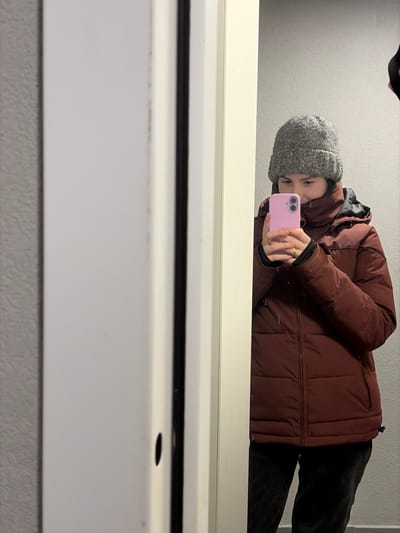 The scene depicts an individual, appearing female, captured in a mirror reflection within an indoor environment in the city of Ковалёво, Russia. The person is shown from mid-thigh up, wearing a grey knit beanie and a high-collared, rust-red or maroon puffer jacket over dark trousers. She holds a light pink smartphone, equipped with dual rear cameras, in both hands at chest level, positioning it to capture her reflection in the mirror. A ring is visible on the ring finger of her left hand. The immediate background behind the reflected person is a plain, light grey wall. The left side of the image prominently features a white door frame or trim, which partially obstructs the view of the reflection. This frame includes a vertical dark strip or seal and a small, dark oval-shaped opening. The wall surface adjacent to the white frame on the far left exhibits a textured, muted grey finish.