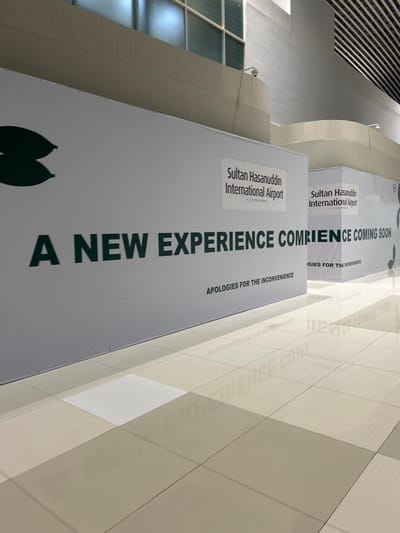 The image shows a large, white banner with dark green text that reads "A NEW EXPERIENCE COMING SOON". Below this, in smaller text, it says "APOLOGIES FOR THE INCONVENIENCE". There are also two smaller signs on the banner, one on the left and one on the right. The sign on the left reads "Sultan Hasanuddin International Airport By Waypoint Airports". The sign on the right is partially obscured but appears to have the same text. The banner is in what appears to be an indoor public space, possibly an airport terminal, with a tiled floor reflecting the banner's text. In the background, there are walls and a ceiling with some architectural details. The overall impression is one of renovation or preparation for a new development at the Sultan Hasanuddin International Airport.