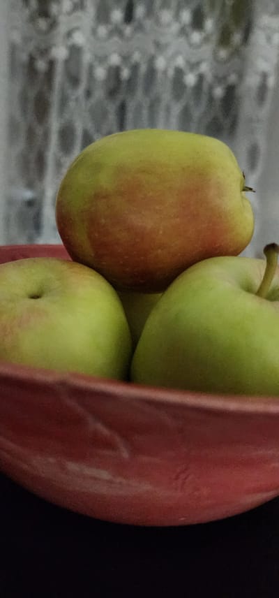A close-up, low-angle shot captures a red bowl filled with apples, set against a blurred background of sheer curtains. The apples, primarily green with hints of red blush, are stacked, with one apple positioned on top of two others. The bowl's rim is visible in the foreground, showing its textured surface. The overall scene is dimly lit, with a shallow depth of field that blurs the background details. The image does not depict any specific identifiable location within Sderot, Israel, nor does it contain any people or visible signs of ongoing activity.