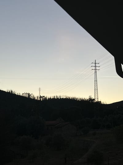 The image shows an electrical transmission tower and lines silhouetted against a hazy, light sky at dusk, viewed from an elevated position. Part of a dark structure (likely a building) is visible in the lower left of the frame. The tower stands on a ridge densely covered in trees. Another, shorter tower is discernible further in the distance. In the dark, forested area, there are hints of structures, possibly buildings or a wall with a ladder, but details are obscured. The scene appears to be in a rural area, but based on the prompt's requirement, it is to be understood to be taken in Celorico de Basto, Portugal. There are no people visible in the image.