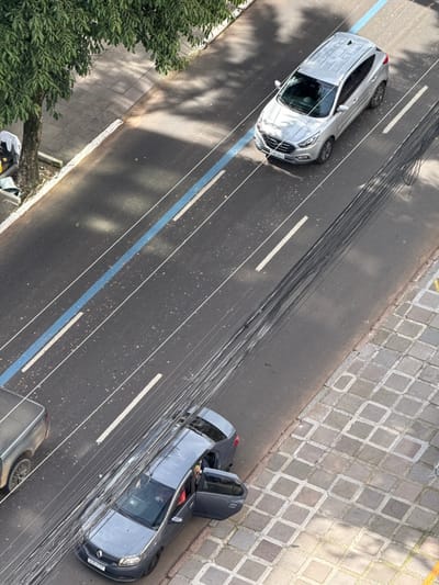 An overhead photograph captures a street scene in Porto Alegre, Brazil, under daylight conditions. The asphalt road features two visible lanes separated by white dashed lines, with an additional solid blue line marking the left lane boundary. On the left side of the road, a concrete sidewalk is visible, bordered by a large tree with green foliage that casts shadows onto the street. Near the tree trunk, white fabric and a yellow object are discernible. In the far-left lane, a light-colored pickup truck is partially visible. Centered in the road, a silver Hyundai Tucson SUV, displaying Brazilian license plate I**B-2405, is in transit or stopped. On the right side of the image, a gray Renault Logan sedan, with Brazilian license plate I**F-3227, is parked or stopped adjacent to a sidewalk paved with square, light-colored tiles arranged in a diagonal pattern. The front driver's side and rear driver's side doors of the Renault Logan are open. A person wearing a light-colored top is visible in the driver's seat, and a partial view of another person's head is seen in the rear through the open door. A bundle of numerous dark utility wires spans diagonally across the middle and right sections of the frame, positioned above the street.