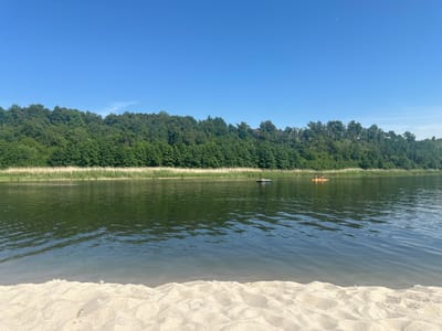 A wide shot captures a tranquil summer scene on a sandy shore overlooking a calm body of water. In the foreground, textured sand forms gentle dunes, leading towards the water's edge. The water itself is a deep green, reflecting the lush trees that line the opposite bank. A dense forest of deciduous trees creates a vibrant green backdrop, stretching across the horizon under a clear, pale blue sky. In the distance on the water, two small rafts or kayaks are visible, each with one or two individuals aboard, suggesting leisurely activity on the water. The overall impression is one of peaceful recreation in a natural setting. The location is identified as Yantarny, Russia.