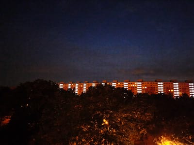 The image depicts a long, multi-story apartment building at dusk or night. The building is illuminated with many brightly lit windows, suggesting that people are inside their homes. The building is orange and has a row of lights across the top. In the foreground, there are dark silhouettes of trees, with some branches and leaves visible. There are also some lights visible within the trees, possibly streetlights or other ambient lighting. The sky above is a dark, deep blue, transitioning to a lighter shade near the horizon, indicating the fading light of day or the approaching night. The overall mood is quiet and serene, with a sense of urban life continuing in the background. The image appears to be taken from a slightly elevated position, looking towards the building and trees. There is no visible text in the image.
