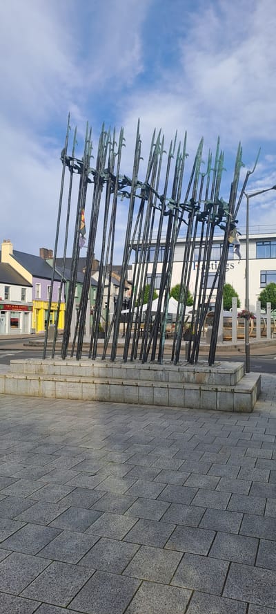 The image shows a public space in Castlebar, Ireland, featuring a sculpture composed of numerous tall, thin metallic poles with pointed tips, arranged in a semi-circular fashion. The sculpture is erected on a tiered stone base, which in turn is situated on a paved area made of grey rectangular tiles. In the background, there are several buildings, some painted in bright colors like yellow and purple, and others in white with prominent signage. A street with parked cars is visible, and beyond that, more buildings and trees. The sky above is blue with scattered clouds. The overall scene depicts an urban environment with a prominent artistic installation.