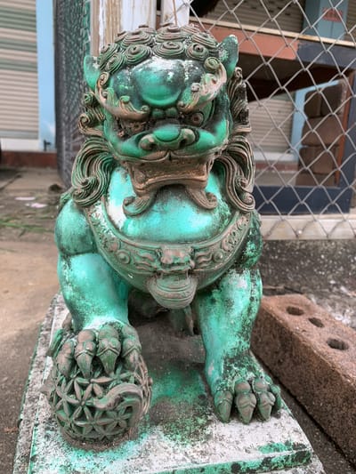 A weathered, green-patinated statue of a Foo dog, likely a guardian lion, stands on a concrete platform outdoors. The statue is detailed, with a mane, ornate collar, and one paw resting on a decorative sphere. The green patina is chipped and weathered, revealing underlying bronze or copper tones in places. The statue is situated in what appears to be a back alley or outdoor storage area. In the background, a chain-link fence, a corrugated metal structure, and a brick are visible. The ground is paved with concrete, and some debris is scattered around. The lighting suggests it is daytime. The location is identified as Ban San Ton Kok, Thailand. No people are present in the image.