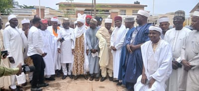 The media file shows a group of approximately fifteen men, most of whom are wearing traditional Islamic attire. They are standing in what appears to be an outdoor setting, possibly in front of a building with a beige facade and a red-tiled roof. The sky is overcast, suggesting a cloudy day.

The men are dressed in various styles of white and light-colored robes (known as agbadas or boubous) and some are wearing kufi caps or turbans. One man in the center has a distinct, richly patterned robe in yellow, white, and brown. Another man on the far right wears a white robe with intricate embroidery. Several individuals are holding mobile phones, and two people in the foreground are holding microphones, suggesting a media event or gathering.

The expressions on the faces of the men are varied. Some are smiling or looking engaged, while others appear more neutral. The overall atmosphere seems to be one of a formal or semi-formal gathering, possibly a religious ceremony, a meeting, or a press event, given the presence of media equipment.

There are no discernible signs or text visible in the image. The location context suggests this event is taking place in Sabon Gari North, Nigeria. The time of day is not clearly indicated but appears to be daytime due to the ambient light.