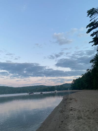 The image depicts a serene natural landscape at the shoreline of a lake in Duhamel, Canada. The sky is a pale blue with scattered clouds, tinged with pink hues near the horizon, suggesting either dawn or dusk. A gentle mist hovers over the calm water, reflecting the sky and the distant tree-covered hills. Several boats are visible on the lake, some anchored, others potentially moving.
The foreground features a sandy beach, with a distinct line where the sand meets the water. A thin white pole stands upright on the beach, likely marking a boundary or a path. The right side of the image is dominated by a dense line of dark green coniferous trees, which extend upwards and frame the sky. The overall atmosphere is tranquil and peaceful, characteristic of a natural lakeside environment.