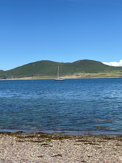 The image depicts a serene coastal landscape under a clear blue sky. In the foreground, a pebble beach is visible, with a gentle tide lapping at the shore, carrying strands of seaweed. The water is a deep, calming blue, with slight ripples on its surface. Further out in the sea, a white sailboat with a tall mast is anchored. In the middle ground, a small, circular pink object, possibly a buoy or a life raft, floats on the water. Dominating the background is a large, green, hilly landscape, its slopes textured with patches of grass and scattered structures, suggesting it is either a mountain or a very large hill. The overall impression is one of natural beauty and tranquility.