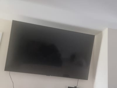 The image shows a large, black television screen mounted on a white wall in a room. The television is turned off, as indicated by the dark screen with some faint reflections. A thin black cable hangs down from the bottom left of the television, disappearing out of frame. The wall appears to be painted a light color, possibly off-white or cream. To the right of the television, a portion of a white corner of the room is visible, suggesting a wall meeting another wall or a structural element. The lighting in the room is diffused, casting soft shadows on the wall. No people are present in the frame. The overall scene is static and appears to be an interior shot of a living space or entertainment area. The image does not provide any specific visual cues to indicate its location within Tulach Mhór, Ireland.
