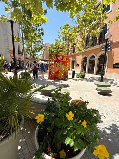 The image captures an outdoor shopping area in the city of La Roca del Vallès, Spain, specifically at the La Roca Village outlet mall. The scene is illuminated by bright sunlight, casting distinct shadows on the paved ground. In the foreground, lush green plants with bright yellow flowers in white planters are visible. A large palm-like plant is also in a white planter to the left. The middle ground features a street lined with shops. The architecture is characterized by ochre-colored buildings with arched windows and balconies. A prominent display of colorful floral artwork, appearing as a large rectangular structure, stands in the center of the walkway. Several people are seen walking along the street. To the left, a black lamppost with a white globe light is positioned. In the background, more trees with green foliage are scattered throughout the area. Several small, round, green tables are placed on the walkway. The overall atmosphere suggests a vibrant and bustling commercial district on a clear day.