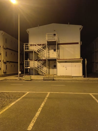 This is a nighttime, eye-level, medium shot photograph of a modern, multi-story building with an external metal staircase. The building is white with a dark red stripe across its upper half. The staircase is also white and leads to multiple levels of the building. At the base of the staircase is a small, white, single-story extension with double doors. A simple wooden bench is placed in front of the extension. To the left of the staircase, a large potted planter with striped flowers sits on a gravel area. A tall street lamp with a bright light source is prominent on the left side of the image, casting a strong yellow glow. In the foreground, a paved road or parking lot is visible, marked with white lines. The area appears to be an urban or industrial setting within Қосшағыл ауылдық округі, Kazakhstan, given the architectural style, which is often seen in modular or prefabricated structures for temporary housing or industrial facilities. The scene is illuminated by artificial lighting, indicating it is after dark. There are no people visible in the image. The environment is clean and well-maintained.
