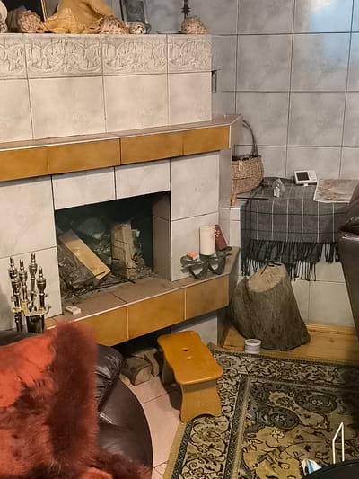 The image shows a living room in Žiaukos, Lithuania, dominated by a fireplace. The fireplace is made of white tiles with a decorative border and a brown wooden mantelpiece. The hearth contains unlit logs, some of which are partially covered by other debris. To the left of the fireplace, there is a set of brass fireplace tools. In front of the fireplace, a dark leather armchair with a reddish fur throw is visible.
To the right of the fireplace, a large, rough-hewn tree stump serves as a decorative element. Behind the stump, a wooden stool is placed on a patterned rug. A woven basket containing what appears to be more logs or branches sits on a gray, fringed blanket draped over a surface, next to a small white electronic device and a stack of papers. A single candle in a wrought-iron holder stands on a wooden shelf to the left of the basket. The walls are tiled in a light gray color. The floor is covered by a patterned rug and wooden planks. The scene depicts a cozy, rustic interior.
