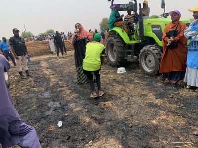 The image shows a gathering of people in an open field in Ringimawa, Nigeria. The field is covered in charred vegetation, indicating a recent fire. A green tractor is parked in the field, with several people standing around it. One woman is speaking into a microphone, while another woman is listening intently. A group of people, including a woman in a green headscarf, are sitting in the tractor's cab, waving. A group of women are standing to the right of the tractor. The woman in front is wearing a red hijab and a brown abaya. She is looking at the woman speaking into the microphone. The other women are wearing blue and white hijabs and abayas. Some people are wearing hats. The overall scene is one of community gathering and activity. The coordinates provided are for Ringimawa, Nigeria.