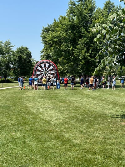 This is a horizontal, full-shot, outdoor photograph taken on a sunny day in Razliv, Bulgaria. The scene depicts a group of people gathered on a grassy lawn in front of a large inflatable dartboard. The dartboard is red and black, with white numbers marking the scoring sections. Several individuals are standing around the dartboard, with some appearing to be participating in a game or observing. A few people are holding what seem to be soft, throwable objects, possibly darts for the inflatable board. The background features lush green trees, suggesting a park or recreational area. The overall atmosphere appears to be casual and social, possibly a team-building event or a community gathering.