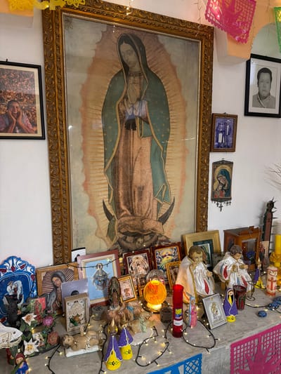 A wall displays a chaotic assemblage of religious iconography and decorative objects. Prominently featured is a large, framed depiction of the Virgin of Guadalupe. Smaller framed pictures, statuettes of baby Jesus figures, religious candles, and string lights cluster densely to its right. Above, a framed portrait, paper decorations, and other religious artifacts contribute to the eclectic, cluttered aesthetic. Below the large painting is another framed picture.