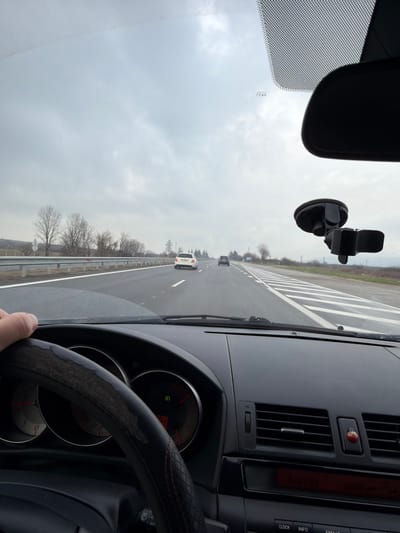 The image depicts a driver's perspective from inside a vehicle traveling on a multi-lane highway, identified as being near the city of Zlatina, Bulgaria. The driver's right hand grips the left side of a dark steering wheel, which is covered with a worn, textured material exhibiting peeling on the upper left and red stitching on the right.
The vehicle's dashboard, dark in color, is visible beneath the windshield, featuring a partially obscured instrument cluster with two circular gauges. A green indicator light and a "0" are visible on the right gauge, while the left displays red markings. To the right of the steering wheel, horizontal air vents are present, with a small red ladybug-shaped ornament attached to one. Below the vents, a segment of the vehicle's infotainment system, including a red digital display and buttons labeled "CLOCK," "INFO," and "FM/AM," is visible. A black rearview mirror and a black, empty suction-cup phone mount are affixed to the upper right area of the windshield.
The highway ahead appears wet, with clear white solid and dashed lane markings. To the right of the observer's lane, a broad section of white diagonal stripes marks a merging area or shoulder. Two other vehicles are visible ahead in adjacent lanes: a white sedan in the middle distance and a darker sedan further ahead. A metal guardrail runs along the left side of the highway. The surrounding landscape consists of bare deciduous trees and sparse vegetation on both sides, suggesting a cold season. Utility poles are visible in the distant background on the left. The sky is uniformly overcast and gray.