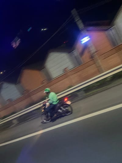 This image captures a night scene in Binh Thuan Ward, Vietnam, with motion blur indicating movement. A person wearing a green helmet and a bright green jacket is riding a motorcycle on a road. The motorcycle has a visible red taillight. In the background, a brick wall borders buildings with white roofs. Above the buildings, a bright blue LED light fixture is illuminated. The scene appears to be an urban or suburban setting at night.