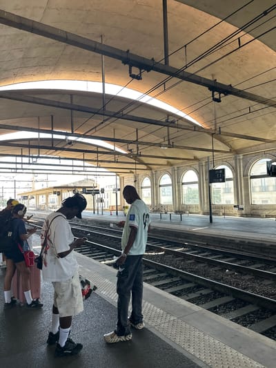 Four individuals are standing on a train platform in Nîmes, France. Two of the individuals in the foreground are looking at their phones. One individual is wearing a white t-shirt and light-colored shorts, while the other is wearing a light blue t-shirt and dark pants. Another individual, seen from the side, is wearing a dark t-shirt, shorts, and headphones, with a pink suitcase next to them. A fourth individual, wearing a yellow cap, is also visible. The platform is next to train tracks, and beyond the tracks are arched windows of a building. The scene is under a covered train station ceiling.