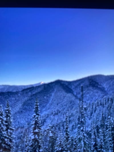 A winter landscape fills the right portion, showing snow-covered mountains and trees. To the left, a bright blue sky suggests a clear day. The contrast between the vibrant sky and the snow-laden terrain creates a striking visual divide. The overall orientation of the image is rotated 90 degrees clockwise.