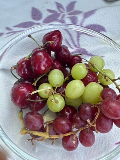 A close-up, overhead shot showcases a glass bowl filled with a vibrant mix of red cherries and green grapes. The fruit is artfully arranged, with some grapes still attached to their delicate stems. The red cherries are plump and glossy, their deep crimson hue catching the light. The green grapes, on the other hand, have a more translucent quality, with a subtle yellow undertone.

The bowl itself is made of clear glass, its curved edges visible and reflecting the surrounding light. It rests on a white textured surface, possibly a tablecloth or placemat, which features a subtle, repeating pattern of purple leaf-like shapes. This pattern adds a touch of understated elegance to the scene.

The lighting suggests it is daytime, with natural light illuminating the fruits and casting gentle shadows. There are no people visible in the image. The overall impression is one of freshness and abundance, perhaps suggesting a healthy snack or a prepared dessert. The location context of Gstadt am Chiemsee, Germany, while not visually evident in the image, adds a touch of European charm to the scene. There is no visible text in the image.