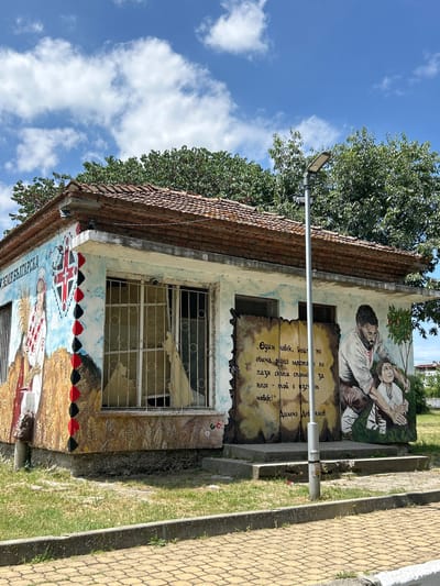 The image shows a mural painted on the exterior of a small, weathered building in Aheloy, Bulgaria. The mural depicts traditional Bulgarian cultural imagery. On the left side, a woman in traditional attire holds a sheaf of wheat, with Bulgarian script above her. A stylized flag design is also present. The central part of the mural features a quote in Bulgarian text written on a parchment-like background, alongside a depiction of a man and a child. The man, with a beard and wearing traditional clothing, is shown placing his hand on the child's head. A small tree with red fruit grows next to them. The building itself has a tiled roof, barred windows, and is situated on a paved area with grass and a sidewalk. A streetlamp stands in front of the mural. The sky above is blue with scattered clouds.