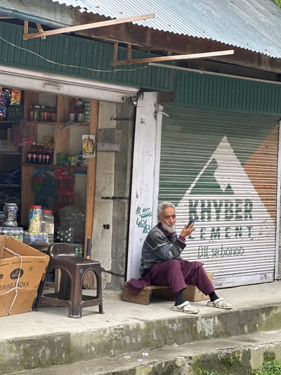 An elderly man with a white beard and grey hair is sitting on a small wooden bench outside a shop in Kullan, India. He is holding a smartphone in his right hand and appears to be looking at it. He is wearing a dark patterned sweater over a maroon shirt and maroon pants, with black socks and white Crocs. The shop has a green corrugated metal facade with a roller shutter that is partially rolled down. A large white and green triangular logo with the word "KHYBER CEMENT" is painted on the shutter, along with the Hindi phrase "Dil se bannao". To the left of the man, there is a brown plastic stool, a cardboard box labeled "PUNJAB TADKA", and shelves stocked with various goods, including bottles of drinks and packaged items. The shop appears to be a small convenience store. The ground in front of the shop is concrete, with some moss and dirt. The overall environment suggests a roadside stall in a town or village.