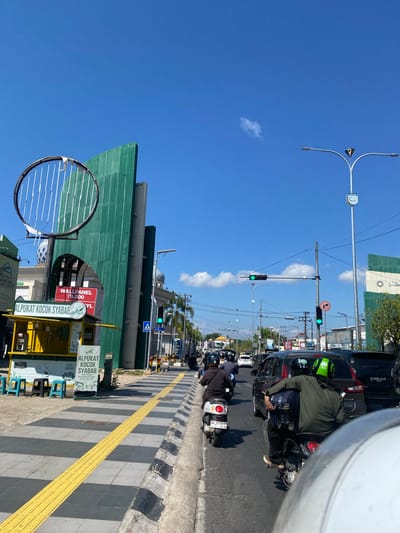 The image depicts a bustling street scene in Jawa, Indonesia, under a clear blue sky. In the foreground, a person is riding on the back of a motorcycle, looking forward at the traffic ahead. They are wearing a green shirt and a helmet with the "Gojek" logo. The motorcycle is in motion, indicating movement through the city.
The street is lined with various establishments, including a bright yellow food stall advertising "ALPUKAT KOCON SYABAB" (Avocado Shake Syabab). Behind the food stall, a large, green, corrugated metal structure stands tall, with a circular, wire-frame sculpture attached to its front. Further down the street, buildings with domed roofs, characteristic of mosques, are visible. Traffic signals, some displaying green lights, indicate the flow of vehicles. Cars and numerous motorcycles are present on the road, suggesting a busy urban environment. The sidewalk features a distinctive yellow tactile paving strip, designed for visually impaired pedestrians. The overall atmosphere is one of daily urban life, with people commuting and engaging in commerce.