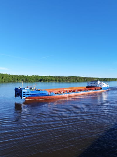 A dry cargo vessel, identified as "ОКСКИЙ-45 И" on its bow and "ОКСКИЙ 45 И" on its stern superstructure, traverses a wide river near Novodevich'e, Russia. The ship features a blue hull, a red-orange anti-fouling bottom, and a white superstructure with blue trim at the stern. Its multiple open cargo holds exhibit an orange-brown interior, indicating either an empty state or the presence of bulk material such as sand or ore. The vessel is oriented diagonally, moving from the left foreground towards the right background, creating a distinct wake and ripples on the dark blue-brown water. The far bank of the river is lined with a dense, unbroken stretch of green deciduous forest under a bright, clear blue sky. A few faint, wispy contrails are visible in the upper atmosphere. A dark, undefined shadow occupies the lower right portion of the image, extending over the water. No individuals are discernible on the vessel or in the surrounding landscape. The scene is illuminated by direct sunlight, indicating daytime.