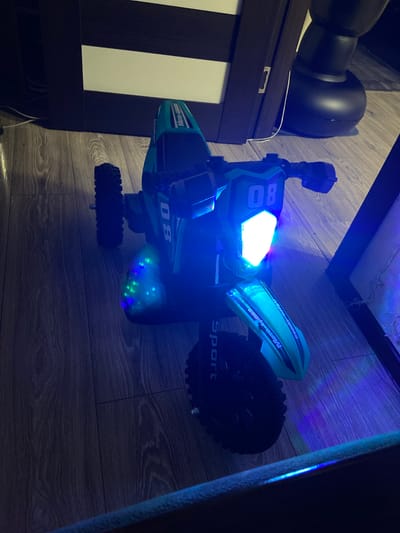 A toy motocross bike with blue and green lights is displayed on a wooden floor. The bike is predominantly teal and black with "Moto-Cross" written on the fenders and "Sport" on the rear wheel guard. The number "08" is visible on the front of the bike, next to a bright white headlight. Several green and blue LED lights are scattered across the underside of the bike, illuminating the floor beneath it. The scene is dimly lit, with the bike's lights being the primary light source, casting blue light onto the surrounding wooden floor and a wall. The background includes a dark brown furniture piece with vertical white stripes and part of a large black object, possibly a speaker or furniture, in the upper left corner. The overall impression is that of a staged display or a close-up product shot in a domestic setting in Beloretsk, Russia. There is no indication of time of day or weather.