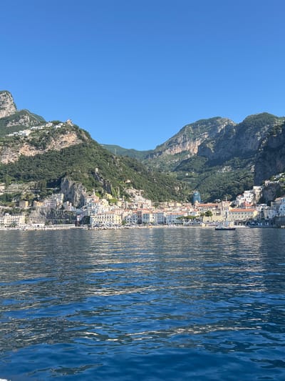 The image depicts a scenic view of the coastal town of Pogerola, Italy, as seen from the water. The town is nestled on a steep, verdant mountainside that plunges towards the azure sea. Buildings with white and pastel facades are stacked precariously along the slopes, interspersed with lush green foliage and rocky outcrops. A clear, bright blue sky dominates the upper portion of the image. The sea in the foreground is a deep blue, with gentle ripples reflecting the sunlight. A breakwater constructed of large rocks lines the edge of the town where it meets the water. In the distance, more of the mountainous coastline is visible, with trees clinging to its steep inclines. There are no people visible in the scene.