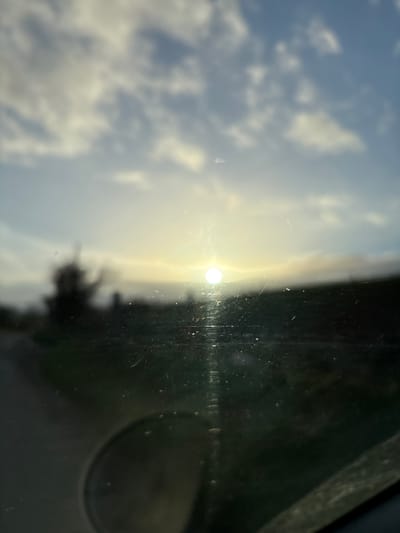 The image depicts an out-of-focus, low-light scene observed through a dirty or scratched glass surface, likely a vehicle windshield, in Shelve, United Kingdom. A bright, circular sun is prominently positioned near the horizon in the left-center, emitting strong glare and vertical light streaks across the viewing surface. A faint halo or lens flare surrounds the sun. The sky above consists of blurry blue with scattered white and grey clouds. The foreground and midground are indistinct, showing a dark, blurry landmass on the right and a suggestion of a road or path leading off to the left, alongside a dark, silhouetted tree-like shape. In the lower-left quadrant, a circular reflection or object, possibly a side mirror, is visible. The bottom edge reveals an indistinct dark structure, likely part of a vehicle's interior. No individuals are present. The environmental conditions suggest either sunrise or sunset.