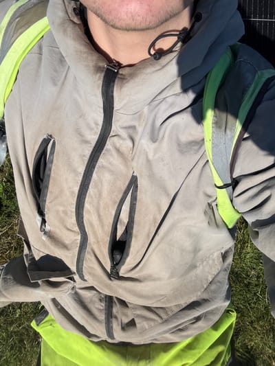 A first-person view captured from above details the upper torso and lower facial area of an individual. The subject wears a light grey hooded jacket, soiled with dark smudges and dirt, featuring a central black zipper and two partially open zipped chest pockets. A neon yellow-green reflective strap with a central grey reflective strip is visible over the right shoulder area, consistent with safety wear. Below the jacket, a portion of a neon yellow-green garment is visible. The individual exhibits stubble on their chin and jaw. The immediate environment includes green grass, indicating an outdoor setting. A dark, gridded rectangular object, consistent with a solar panel, is partially visible in the upper right background. The scene is illuminated by bright sunlight, casting sharp shadows. The specific location within Southampton, United Kingdom, is not discernible from the visual evidence.