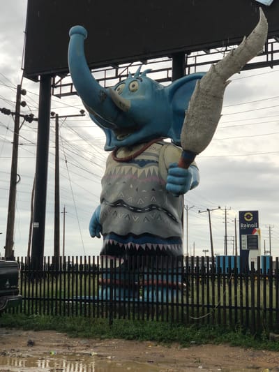 A large, blue, cartoon-like elephant statue stands on a foundation, holding a gray, bushy object that resembles a tree branch or a large duster. The elephant has a long trunk raised upwards, large ears, and wide eyes. It is wearing a patterned vest with geometric designs in gray, pink, and blue. The statue is situated in front of a black metal fence and behind it, there are numerous utility poles and overhead wires, suggesting an urban or semi-urban setting. To the right of the statue, a tall, dark blue and white sign with the word "Rainoil" and a circular logo is visible, indicating a gas station. The ground in the foreground is muddy with puddles of water, and some green vegetation grows along the fence line. The sky is overcast, with a cloudy, gray appearance. The overall scene is that of an outdoor monument or advertisement, likely located in the city of Magado, Nigeria.